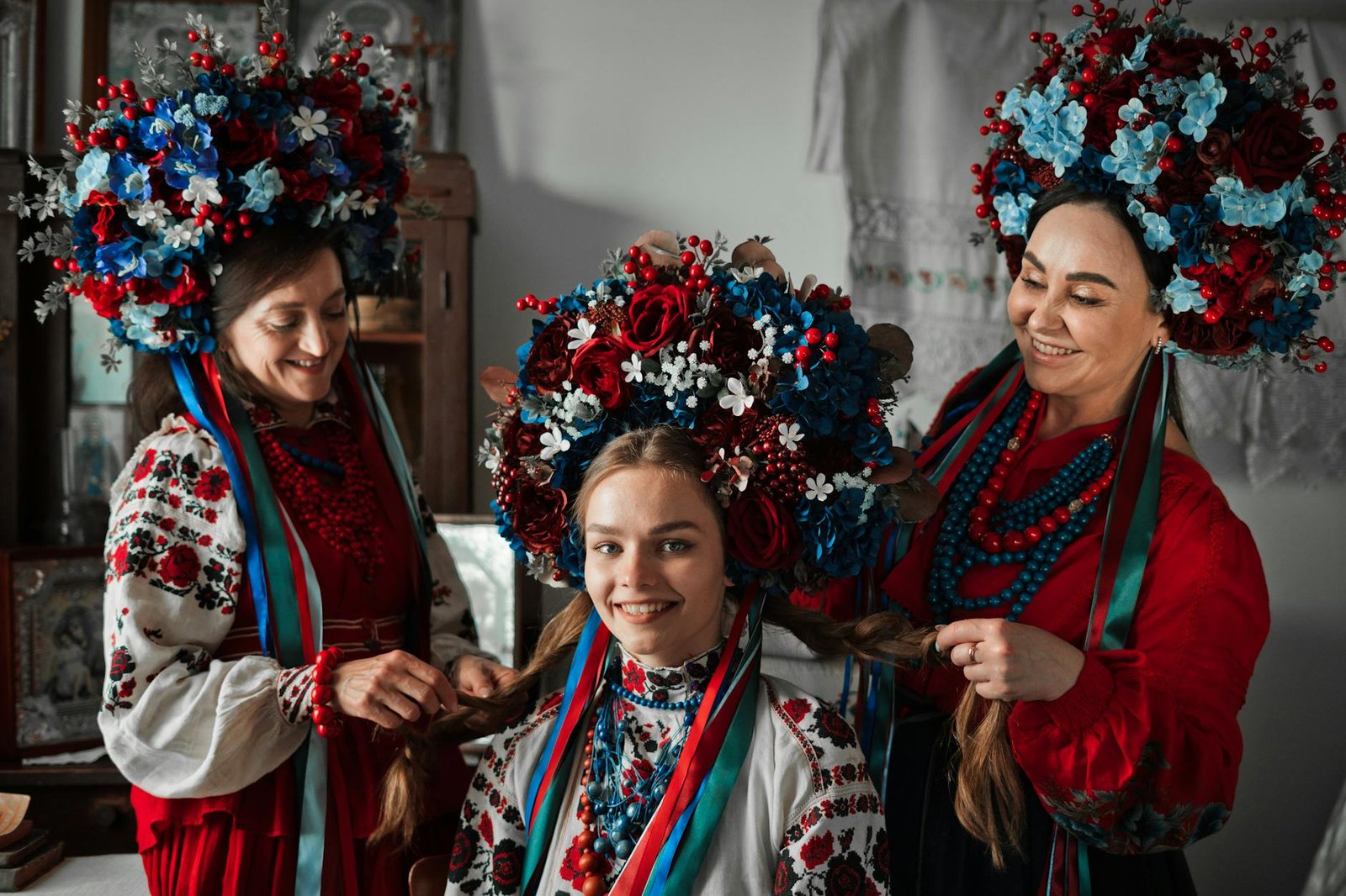Three women in vibrant traditional Ukrainian attire with intricate floral headdresses indoors.