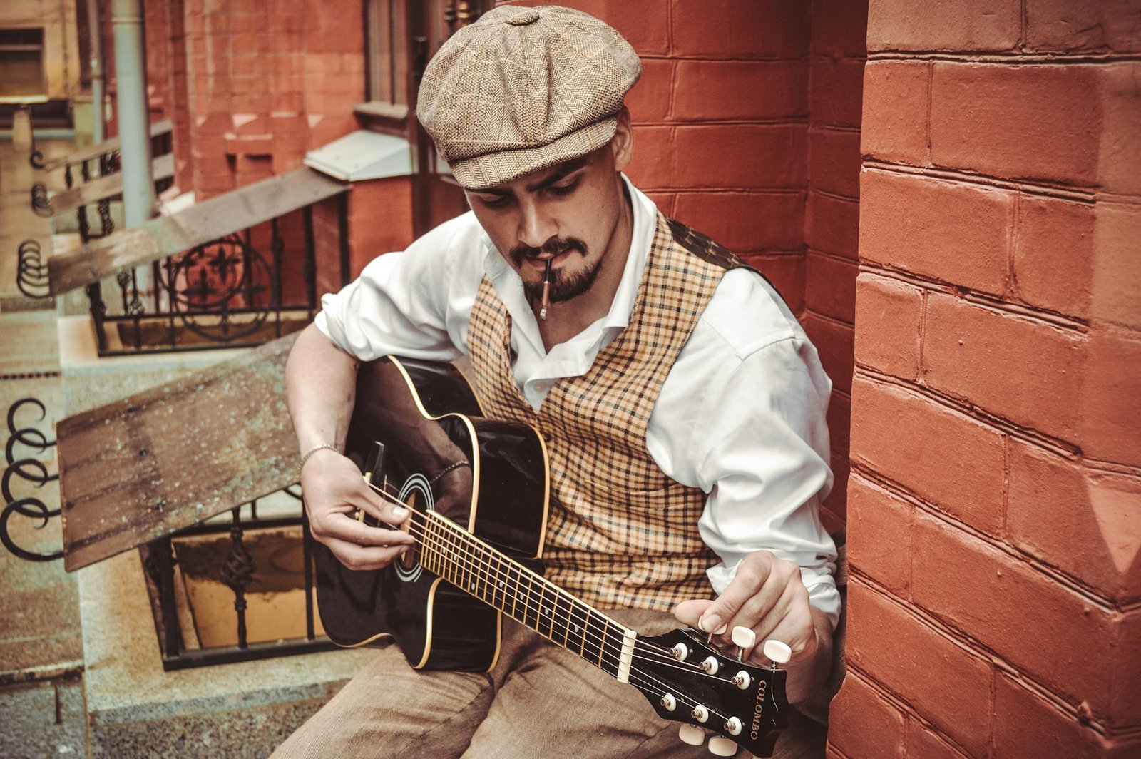 Focused male musician in cap and cigarette wearing white shirt sitting near brick house while tuning guitar in city district