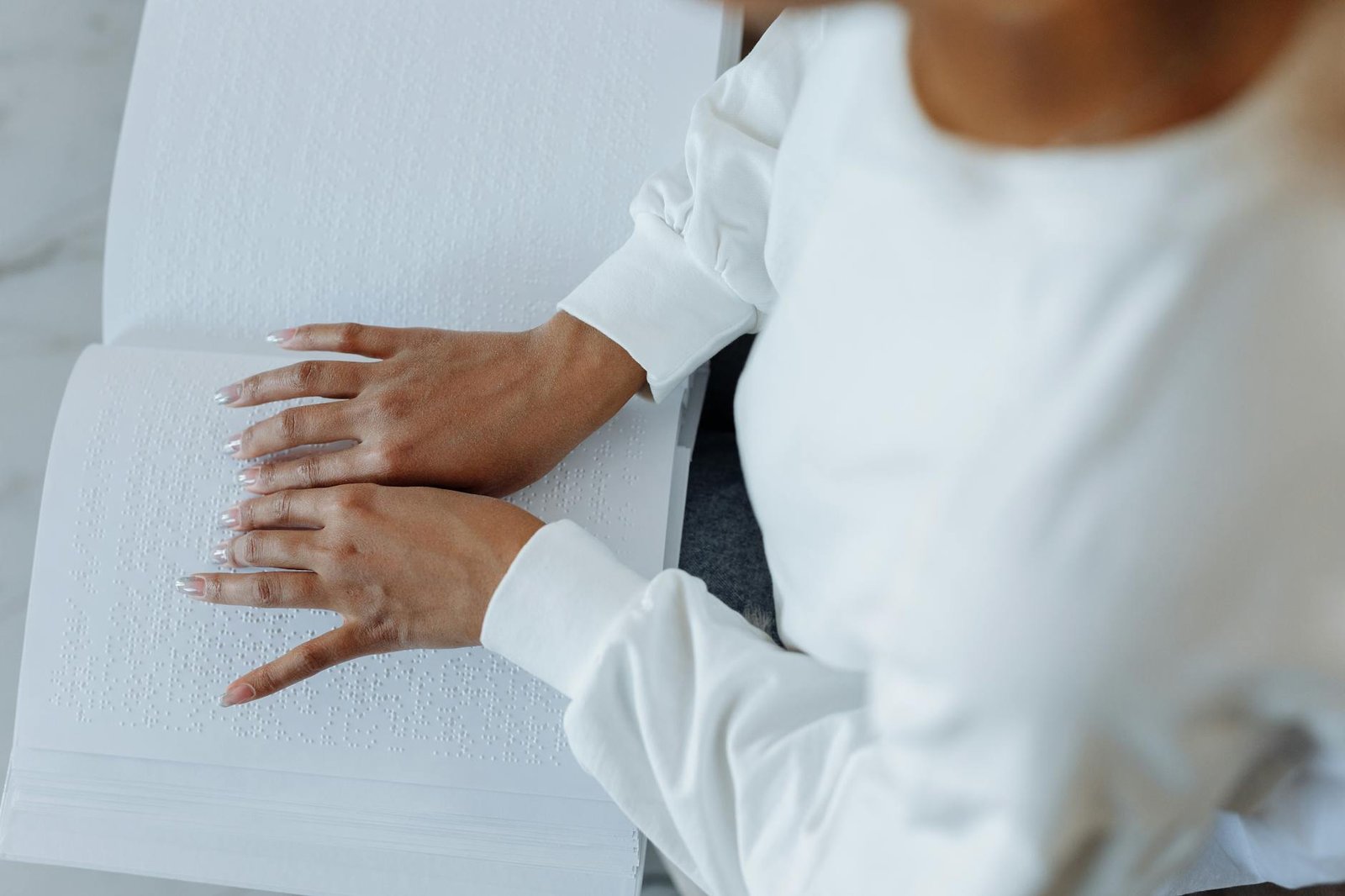Person using hands to read Braille text in an open book while wearing a white sweater.