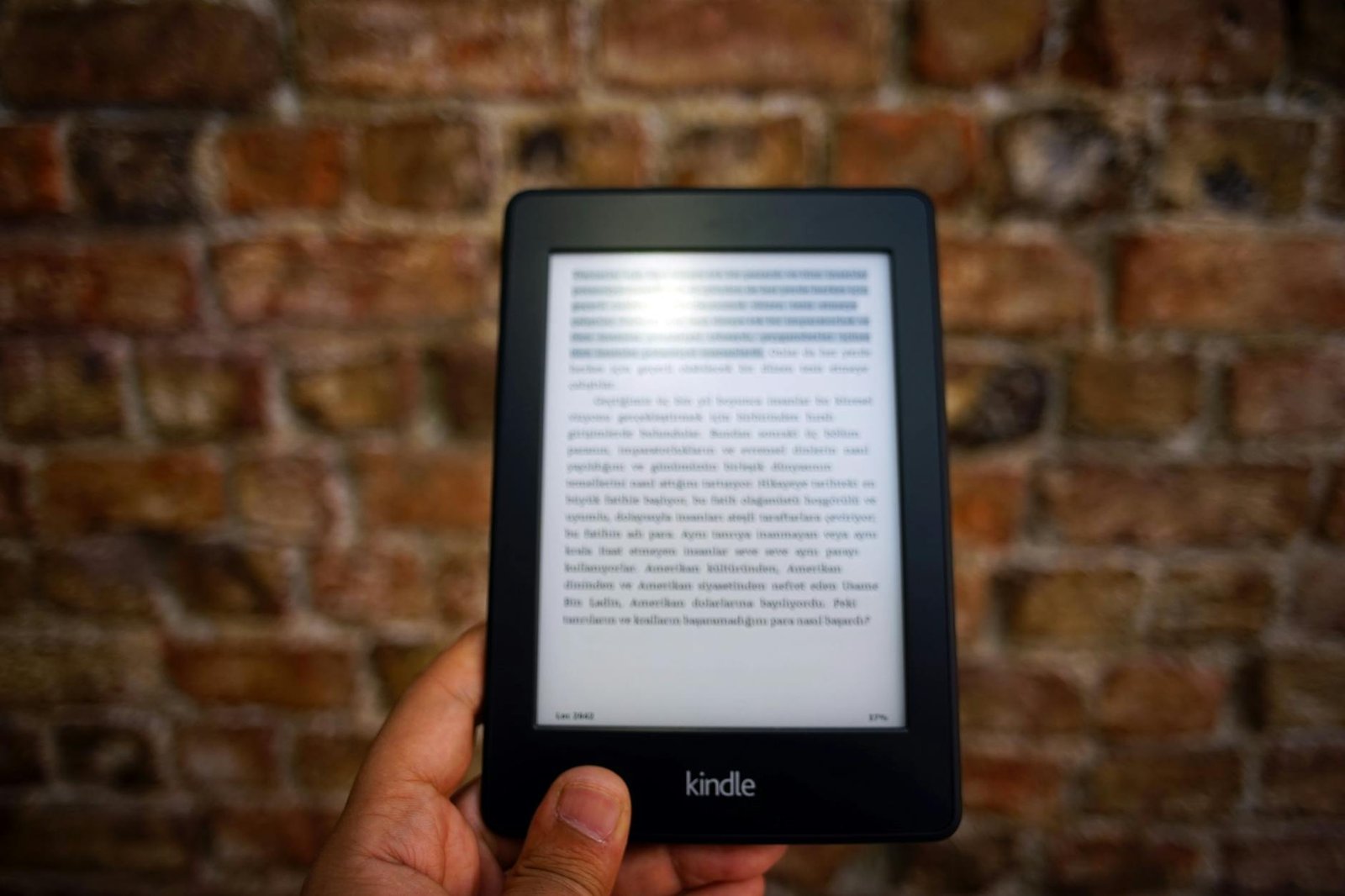 Close-up of a Kindle e-reader held by a person, showcasing a digital reading device against a textured brick wall backdrop.