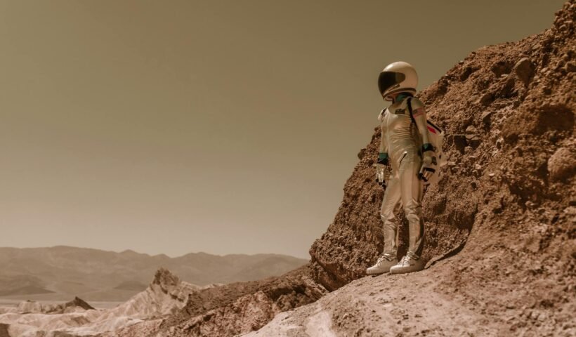 A lone astronaut in a spacesuit stands on a rugged, rocky terrain resembling an alien planet.