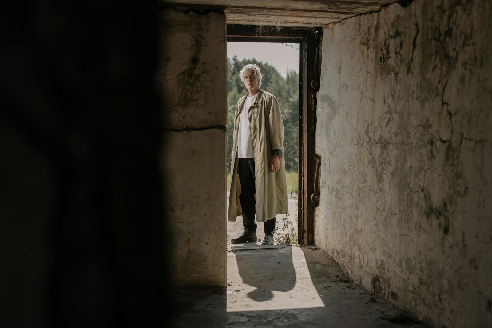 A senior man in a trench coat stands at the entrance of an abandoned building, casting a shadow.