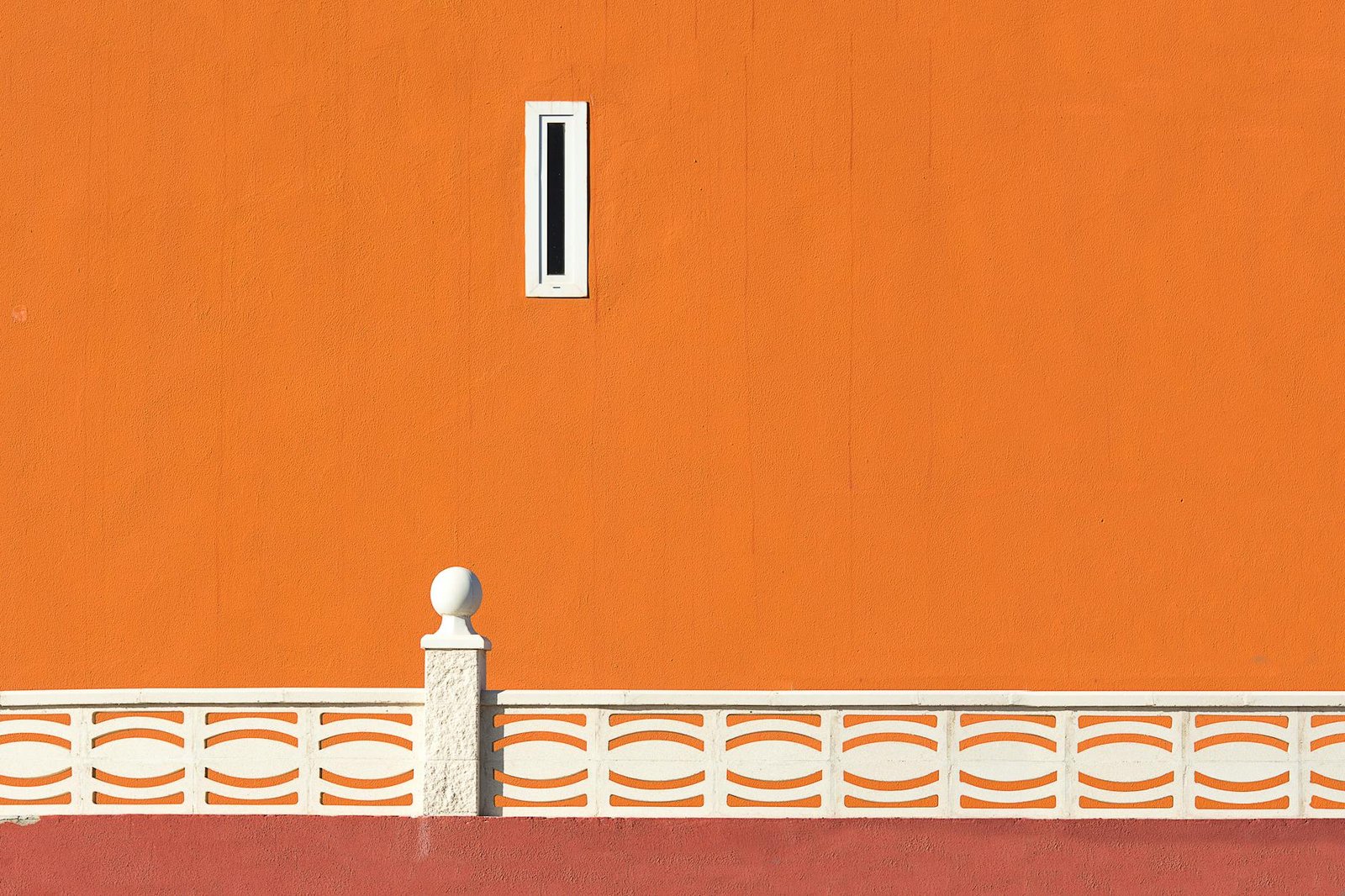 Minimalist orange wall with white patterned railing and a small window in urban setting.