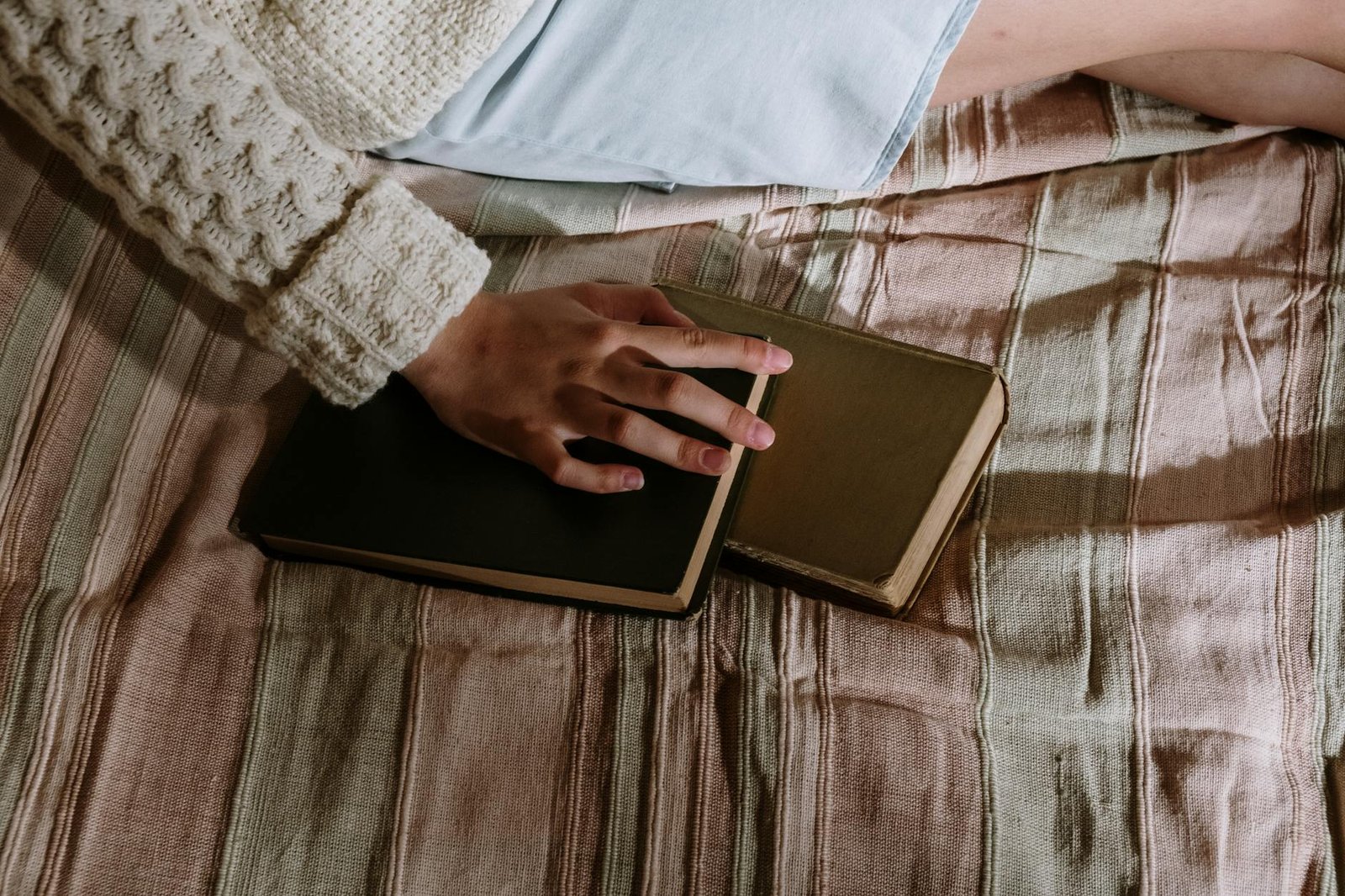 A cozy scene of a person in a wool sweater holding a hardcover book on a bed.
