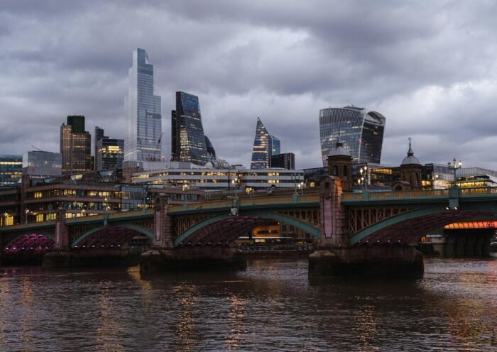 Bridge with glowing lights crossing calm Thames river located against contemporary famous multistory office buildings in London on evening time