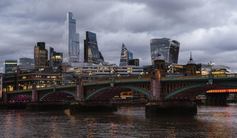 Bridge with glowing lights crossing calm Thames river located against contemporary famous multistory office buildings in London on evening time