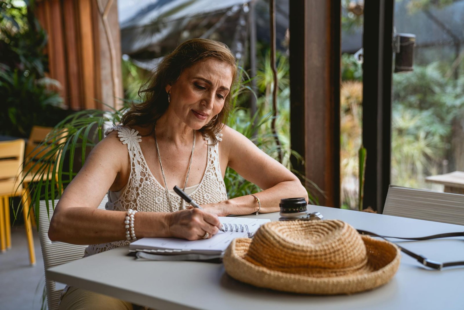 Elderly woman sitting at a cafe table, writing in a notebook with a straw hat nearby.